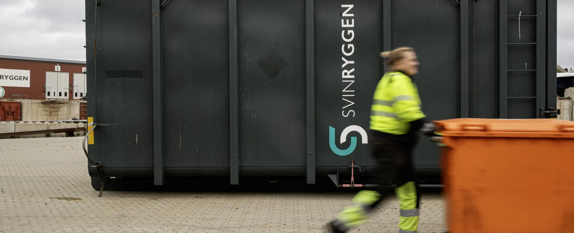 Kontainer med Svinryggens logotyp, en kvinnlig anställd går förbi med ett mindre sopkärl vid Svinryggens kretsloppsanläggning.